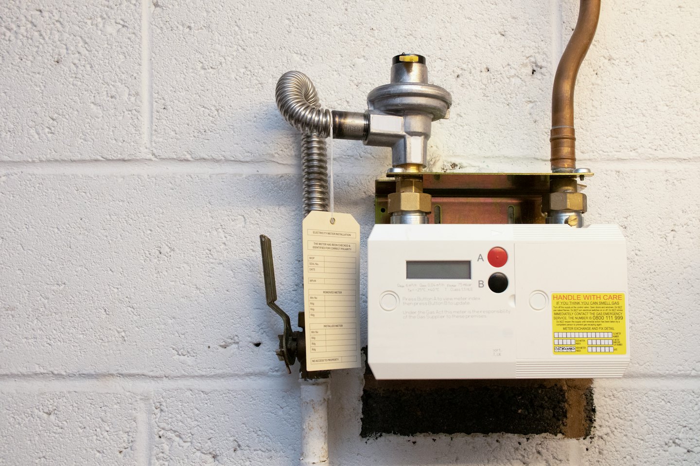 Digital gas meter installation with pipes and warning sticker.